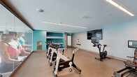 Located at 480 N Orange Ave in the heart of Orlando, FL, this purpose-built spin and cycling studio offers a well-equipped, single-room fitness space designed to support focused, high-intensity workouts. The studio's interior features a clean, modern aesthetic with light gray walls accented by a bold teal feature wall housing open shelving stocked with medicine balls, stability balls, and other workout accessories. Linear LED lighting runs across the smooth white ceiling, providing bright, even illumination throughout the room. The durable, neutral-toned flooring offers a resilient surface suited for cycling and floor exercises alike. Multiple stationary cycling bikes are arranged across the open floor, including spin bikes and an Echelon rowing machine, providing a versatile cardio offering. A large flat-screen television is mounted on the wall, supporting guided workouts and entertainment during sessions. A full-length glass partition along one wall reflects natural light and creates a sense of expanded space, while also allowing views to an adjacent area. A water fountain is conveniently positioned near the entrance, along with sanitation supplies and a first aid cabinet. The space is well-ventilated with visible HVAC registers throughout. Compact yet thoughtfully outfitted, this studio delivers a professional fitness environment suited for both individual training and group classes.