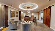 Located at 2432 Albany Ave in West Hartford, CT, this sophisticated lounge encompasses three thoughtfully designed living areas on a single floor. The centerpiece of the space is a dramatic circular tray ceiling with layered cove lighting and a stunning cluster pendant chandelier with teardrop glass shades, casting a warm, inviting glow throughout the main gathering area. The primary lounge features a curved sectional sofa anchored around a round marble-top coffee table, flanked by metallic accent tables and plush accent chairs — all set atop patterned carpeted flooring. Rustic barn-style sliding wood doors provide an eye-catching contrast against the contemporary neutral palette, while dark wood columns with textured metallic wall treatments add architectural depth. A secondary sitting nook offers two streamlined lounge chairs paired with a small side table beneath original artwork, providing an intimate retreat. A third area serves as a media lounge, complete with a wall-mounted flat-screen television set against a warm wood-paneled accent wall, low credenzas, and additional seating. Throughout, curated artwork, layered lighting, and a cohesive warm taupe and earth-tone color scheme create a polished, welcoming ambiance ideal for comfortable gathering and relaxation.