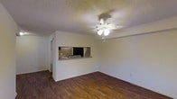 This single-floor space offers a straightforward, functional layout centered around an open living area. The room features dark hardwood-style flooring that contrasts cleanly with the light-colored walls, creating a balanced, neutral aesthetic throughout. A ceiling fan with an integrated light fixture serves as the focal point overhead, providing both ventilation and ambient lighting. The ceiling features a textured popcorn finish, a classic detail that adds subtle depth to the space.

A defining architectural feature is the pass-through window cut into the wall, which connects the living area to the kitchen. This opening is framed with a small countertop ledge, ideal for casual dining or serving, and allows natural sightlines between the two spaces. The kitchen, visible through the pass-through, appears compact and functional. A doorway to the left hints at additional rooms beyond the main living space.

The overall ambiance is clean and understated, with neutral tones and simple finishes that offer a versatile backdrop for a variety of interior styles. The open feel of the main living area, combined with practical features like the ceiling fan and pass-through, makes this a highly functional single-floor space.