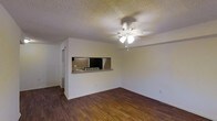 This single-floor space offers a straightforward, functional layout centered around an open living area. The room features dark hardwood-style flooring that contrasts cleanly with the light-colored walls, creating a balanced, neutral aesthetic throughout. A ceiling fan with an integrated light fixture serves as the focal point overhead, providing both ventilation and ambient lighting. The ceiling features a textured popcorn finish, a classic detail that adds subtle depth to the space.

A defining architectural feature is the pass-through window cut into the wall, which connects the living area to the kitchen. This opening is framed with a small countertop ledge, ideal for casual dining or serving, and allows natural sightlines between the two spaces. The kitchen, visible through the pass-through, appears compact and functional. A doorway to the left hints at additional rooms beyond the main living space.

The overall ambiance is clean and understated, with neutral tones and simple finishes that offer a versatile backdrop for a variety of interior styles. The open feel of the main living area, combined with practical features like the ceiling fan and pass-through, makes this a highly functional single-floor space.