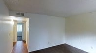 This single-floor space presents a straightforward, functional layout centered around a main living area characterized by neutral white walls and a textured ceiling overhead. The room features dark carpeting that contrasts with the bright, clean wall surfaces, creating a simple yet practical environment. A ceiling vent indicates a central HVAC system for climate control throughout the unit. An open doorway leads from the main living area into a hallway, which connects to additional rooms beyond. The hallway transition reveals hardwood or wood-style flooring, distinguishing it from the carpeted living space. Natural light filters through what appears to be a window at the far end of the hallway, contributing brightness to the interior. The overall layout appears compact and efficiently organized across a single level, with a clear flow between the main living space and adjoining rooms. The minimalist design and neutral color palette provide a blank canvas that can accommodate a variety of furnishing and décor styles.