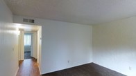 This single-floor space presents a straightforward, functional layout centered around a main living area characterized by neutral white walls and a textured ceiling overhead. The room features dark carpeting that contrasts with the bright, clean wall surfaces, creating a simple yet practical environment. A ceiling vent indicates a central HVAC system for climate control throughout the unit. An open doorway leads from the main living area into a hallway, which connects to additional rooms beyond. The hallway transition reveals hardwood or wood-style flooring, distinguishing it from the carpeted living space. Natural light filters through what appears to be a window at the far end of the hallway, contributing brightness to the interior. The overall layout appears compact and efficiently organized across a single level, with a clear flow between the main living space and adjoining rooms. The minimalist design and neutral color palette provide a blank canvas that can accommodate a variety of furnishing and décor styles.