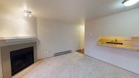 This single-floor space centers around a comfortable living area finished with wall-to-wall carpet in a neutral tone, creating a warm and understated foundation throughout the room. The walls are painted a clean, off-white hue, contributing to a bright and open atmosphere. A fireplace with a simple mantel is positioned along one wall, serving as a natural focal point and adding a functional yet classic design element to the living space. Baseboard heating runs along the lower portion of the main wall, ensuring efficient climate control throughout the room. A pass-through window with a wooden counter is situated on the adjacent wall, providing a convenient connection to a neighboring area — likely a kitchen — and enhancing the overall flow and accessibility of the layout. The transition from carpeted flooring in the living area to hardwood or laminate flooring visible beyond the pass-through adds a subtle contrast in materials. The space is well-lit with ceiling-mounted lighting, and the clean, simple design makes it highly versatile and easy to personalize with furnishings and décor.