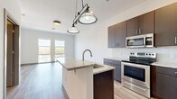 This thoughtfully designed one-bedroom, one-bathroom residence at 383-389 Main St offers a well-utilized single-floor layout with contemporary finishes throughout. Light-toned hardwood-style flooring runs continuously across the living areas, creating a cohesive and clean aesthetic.

The kitchen is a standout feature, equipped with dark espresso cabinetry, granite countertops, a subway tile backsplash, and a full suite of stainless steel appliances including a refrigerator, range, microwave, and dishwasher. A central island with an undermount sink and pendant lighting serves as both a functional workspace and a casual dining area, opening directly to the living space.

The spacious primary bedroom receives generous natural light through large windows and offers direct access to a private balcony, where sweeping views of the surrounding neighborhood can be enjoyed. A second set of dual windows further enhances the bright, airy feel of the room.

The bathroom is finished with large-format floor tiles, a dark vanity with a white countertop, and a full tub-shower combination clad in classic white subway tile. A utility room provides added practicality. Overall, the unit presents a modern, functional living environment with quality finishes and a well-connected floor plan.