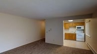 This single-floor space features a straightforward, functional layout with a generously sized living area that flows openly toward a separate kitchen. The living room is finished with wall-to-wall carpet in a neutral brown tone, contributing to a quiet, understated atmosphere. The walls are painted in a clean, light gray-white palette throughout, lending a bright and airy feel to the interior despite the absence of visible windows in the main living space. A thermostat panel is mounted on the living room wall, and standard electrical outlets are positioned at regular intervals.

The kitchen is accessible through a defined opening and contrasts the carpeted living area with hard tile flooring in a classic pattern. Warm honey-toned wood cabinetry lines both the upper and lower sections of the kitchen, providing ample storage space. A white refrigerator is visible alongside a sink with a standard faucet, positioned beneath the upper cabinets. Overhead lighting in the kitchen offers functional illumination. The transition between the two spaces is clearly delineated by the flooring change, making the layout easy to navigate. Overall, the space presents a clean, utilitarian design suited for everyday living.