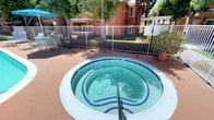 This outdoor recreational area centers around a generously sized swimming pool with a smooth, curved design and clear turquoise water, complemented by an adjacent circular spa featuring blue tile detailing and built-in jets. The pool deck is finished in a warm terracotta-toned surface that surrounds both water features, providing ample space for lounging and socializing. Seating throughout the area includes padded chaise lounges and patio chairs and tables, many shaded by vibrant blue market umbrellas that offer relief from the sun. The perimeter of the pool area is enclosed by a white metal security fence with a self-closing gate, ensuring a safe and controlled environment. An outdoor shower station is conveniently positioned near the pool house entrance, which features white French doors with slatted shutters providing access to restroom or utility facilities. Mature trees lining the property create a natural canopy, adding shade and greenery throughout the space. The surrounding multi-story buildings, constructed of brick and stucco with covered balconies, frame the courtyard and contribute to a sense of privacy. The overall ambiance is bright, resort-like, and well-maintained, making it an inviting outdoor retreat.