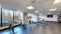 Located at 779 Eves Dr in Hillsborough, NJ, this well-equipped fitness center offers a comprehensive workout environment within a single, open-plan space. The room features durable speckled rubber flooring throughout, providing a safe and resilient surface suited for a wide range of exercises. A drop ceiling with recessed panel lighting ensures the space is consistently well-lit, complemented by large windows along multiple walls that invite ample natural light and offer pleasant views of the surrounding wooded landscape.

The cardio zone is stocked with an array of machines including treadmills, ellipticals, and stationary spin bikes, while a wall-mounted flat-screen television keeps users entertained during workouts. The strength training area is equally well-appointed, featuring a multi-functional cable machine, adjustable weight bench, TRX suspension training system, and a dedicated rack holding kettlebells, medicine balls, and free weights. A storage area with louvered closet doors provides additional organizational space.

Safety features include clearly marked emergency exit signage, a fire extinguisher, and a smoke detector. A wall-mounted intercom panel and a water fountain add convenience. The fitness center's clean white walls, dark accent trim, and open layout create a bright, functional, and motivating atmosphere for residents seeking a professional-grade exercise facility.