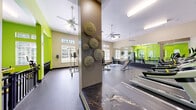 Located at 11100 Beckstone Way in Raleigh, NC, this fitness center offers a well-appointed, single-floor workout facility designed to support a wide range of exercise needs. The space is immediately distinguished by its bold lime green accent walls, which contrast energetically against neutral beige tones and create a vibrant, motivating atmosphere throughout.

The layout is thoughtfully organized into distinct workout zones. A dedicated cardio area houses multiple treadmills and stationary bikes, while a strength training section features cable machines, weight benches, barbells, and a power rack. Yellow-accented free-standing exercise equipment adds further variety for functional training. Durable rubber flooring spans the entire space, providing both safety and comfort underfoot.

Full-length mirrors line one wall, visually expanding the room and allowing users to monitor their form during workouts. Ceiling fans are distributed throughout, ensuring consistent air circulation. Natural light filters in through multiple windows fitted with blinds, complemented by recessed and fluorescent overhead lighting. A wall-mounted flat-screen television adds an entertainment element to the cardio zone.

Decorative spherical wall art on a central column adds a unique design touch, balancing the functional aesthetic with visual interest. The high ceilings further enhance the sense of openness, making this fitness center a comprehensive and inviting space for exercise.