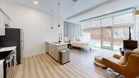 Located at 888 Delaware Ave in Buffalo, NY, this thoughtfully designed studio offers a smart, open-concept layout that maximizes every square foot. Light hardwood-style flooring flows throughout, complementing the clean white walls and contemporary finishes that give the space a bright, modern feel.

The main living area seamlessly integrates the sleeping and lounge zones, anchored by large floor-to-ceiling windows that flood the room with natural light and offer views of the surrounding streetscape. A murphy-style bed with wall-mounted lighting provides flexibility, while space for a desk, seating, and a dresser makes the area highly functional.

The full kitchen is a standout feature, equipped with gray shaker cabinetry, stainless steel appliances including a range, microwave, and refrigerator, and a marble-look countertop peninsula with a double sink and elegant pendant lighting with gold accents. A separate kitchen area accessible via sliding barn-style doors adds a dedicated culinary zone.

The bathroom is clean and well-appointed, featuring a gray vanity with a marble-look countertop, a large mirror with modern lighting fixtures, and a glass-enclosed walk-in shower with subway tile surround and a rain showerhead. The overall ambiance is polished and move-in ready, ideal for efficient urban living.