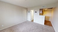 This single-floor studio offers a practical and straightforward layout designed for efficient use of space. The main living area is finished with neutral-toned carpeting and features vertical wood panel accents along one wall, adding a subtle textural element to the room. A textured ceiling runs throughout, and a window at the entry side of the living space allows natural light to filter in. The living area flows toward a partially divided kitchen section, separated by a low white partition wall. The kitchen is equipped with warm-toned wood cabinetry mounted overhead, providing ample storage, and transitions to wood-look flooring that distinguishes it from the carpeted living space. HVAC vents are integrated into the ceiling for year-round climate control. A doorway from the main living area leads to the bathroom, which features durable wood-look plank flooring, a toilet, and a closeted storage area. The overall color palette throughout the studio is light and neutral, with beige and white tones creating a clean, cohesive feel. The space is straightforward in its design, offering a functional environment well-suited for single occupancy.