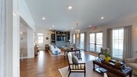 This single-story clubhouse offers a thoughtfully designed communal gathering space that blends classic and coastal-inspired aesthetics. The main living area is anchored by rich, dark hardwood floors that extend throughout, providing a warm contrast to the light, neutral-toned walls and bright white ceiling fitted with recessed lighting. A elegant lantern-style chandelier adds a refined focal point overhead.

A built-in media center finished in a muted blue-gray tone serves as the room's architectural centerpiece, housing a large flat-screen television and open shelving for decorative displays. The seating arrangement features a mix of upholstered sofas and spindle-back accent chairs layered over area rugs, creating distinct yet open conversation areas throughout the space.

Natural light flows generously into the room through multiple sets of French doors along the exterior wall, offering views of the surrounding community and providing easy indoor-outdoor access. Decorative wall sconces and table lamps complement the ambient lighting, enhancing the inviting atmosphere. The overall design communicates a relaxed yet polished environment well-suited for community gatherings and everyday socializing.