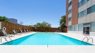 Located at 6303 Indian School Rd NE in Albuquerque, New Mexico, this single-level outdoor amenity space centers around a generously sized swimming pool that serves as its primary feature. The pool area is defined by clear, turquoise water and a broad concrete pool deck finished with decorative tilework at the entry point, including a distinctive sun-burst pattern that adds a Southwestern aesthetic touch. A row of lounge chairs lines one side of the pool along a tall wooden privacy fence, providing a comfortable area for sunbathing beneath Albuquerque's characteristically bright blue skies.

Access to the pool area is through secured glass double doors, underscoring the private nature of the space. The surrounding grounds extend beyond the pool deck to a secondary outdoor patio area featuring a pergola structure with outdoor seating, suitable for shaded gatherings. Mature trees along the fenced perimeter add natural shade and a sense of enclosure. The building's stucco exterior walls and large windows frame the courtyard on one side, while views of adjacent multi-story commercial buildings are visible beyond the fence. The overall atmosphere is open, sun-drenched, and well-suited to the warm Albuquerque climate.