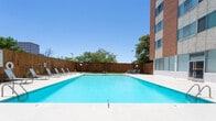 Located at 6303 Indian School Rd NE in Albuquerque, New Mexico, this single-level outdoor amenity space centers around a generously sized swimming pool that serves as its primary feature. The pool area is defined by clear, turquoise water and a broad concrete pool deck finished with decorative tilework at the entry point, including a distinctive sun-burst pattern that adds a Southwestern aesthetic touch. A row of lounge chairs lines one side of the pool along a tall wooden privacy fence, providing a comfortable area for sunbathing beneath Albuquerque's characteristically bright blue skies.

Access to the pool area is through secured glass double doors, underscoring the private nature of the space. The surrounding grounds extend beyond the pool deck to a secondary outdoor patio area featuring a pergola structure with outdoor seating, suitable for shaded gatherings. Mature trees along the fenced perimeter add natural shade and a sense of enclosure. The building's stucco exterior walls and large windows frame the courtyard on one side, while views of adjacent multi-story commercial buildings are visible beyond the fence. The overall atmosphere is open, sun-drenched, and well-suited to the warm Albuquerque climate.