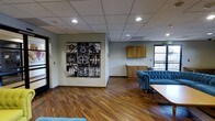 This single-floor space combines modern functionality with creative character, making it a versatile environment suited for a variety of uses. The main area features rich, dark hardwood flooring that anchors the room with warmth and a polished aesthetic. A striking grid-style wall art installation with a bold cinematic film theme serves as a prominent focal point, adding personality and visual interest to the space.

The lounge area is furnished with a tufted blue velvet sofa, lending a touch of elegance and color contrast against the neutral tones of the surrounding walls and drop-panel ceiling. Recessed lighting throughout ensures consistent, even illumination across the space.

Along one wall, a built-in kitchenette provides practical utility, featuring warm wood cabinetry and a countertop with a sink — a convenient amenity for extended use or gatherings. A large window nearby allows natural light to filter in, offering a view to the exterior and contributing to an open, airy atmosphere.

The drop ceiling with integrated recessed lighting gives the space a clean, professional finish. Overall, the design balances a contemporary, creative aesthetic with functional layout, making it well-suited for use as a lounge, meeting area, or collaborative workspace.