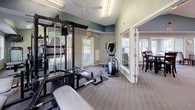This fitness center is thoughtfully designed to support a comprehensive workout experience within a bright and welcoming environment. The space features vaulted ceilings accented by a teal upper wall band, which adds a distinctive visual character while contributing to an open, airy atmosphere. A ceiling fan and fluorescent lighting provide ample illumination throughout.

The workout area is well-appointed with a range of equipment, including a treadmill, elliptical trainer, rowing machine, cable weight machine with a bench, and additional resistance training apparatus. Wall-mounted mirrors expand the sense of space and allow users to monitor their form during exercise. A wall-mounted television adds entertainment options for those on cardio machines.

Multiple large windows line the walls, flooding the room with natural light and offering pleasant views of the surrounding outdoor greenery. A dedicated exterior door provides convenient direct access to the outdoors. Durable carpet flooring covers the workout area, offering cushioning underfoot.

French doors connect the fitness center to an adjacent community lounge and dining area, creating a seamless flow between spaces. The lounge features comfortable seating, track lighting, and an open layout, making it an ideal space to relax before or after a workout. Together, these spaces form a cohesive and functional community amenity area.