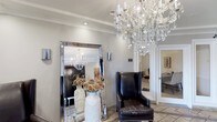 This refined single-floor space exudes an air of timeless elegance from the moment you enter. The reception area immediately captivates with a grand crystal chandelier that casts a warm, luminous glow across the room, setting a luxurious tone throughout. Crown molding frames the ceiling with classic architectural detail, while neutral warm-gray walls provide a sophisticated backdrop that complements the rich interior furnishings.

A large floor-length mirror with a sleek mirrored frame amplifies the sense of space and reflects the chandelier's brilliance, adding depth and grandeur to the entryway. A striking black leather wingback chair with nailhead trim offers a bold, refined seating accent, perfectly balanced by an elegant white ceramic vase filled with warm-toned floral arrangements that add a natural, welcoming touch.

Recessed lighting supplements the chandelier, ensuring the space is both functional and atmospheric. Wall-mounted accent sconces further enhance the layered lighting design. Light-toned tile flooring grounds the space while maintaining a clean, polished aesthetic. A doorway visible in the background hints at additional rooms beyond, suggesting a thoughtfully connected layout. Overall, the space presents a seamlessly curated environment that is both inviting and impressively appointed.