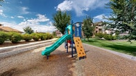 Located at 1600 S Taft Ave in Loveland, Colorado, this outdoor playground and community green space offers a well-appointed recreational environment designed for active outdoor enjoyment. The centerpiece of the space is a vibrant, multi-component play structure featuring a rock climbing wall, a winding slide, and various interactive elements set atop a safety-conscious wood chip surface. A second, larger play structure adds additional slides and play components, providing options for children of varying ages and abilities. Surrounding the play areas, expansive stretches of lush, manicured lawn are framed by mature deciduous trees that provide natural shade and a welcoming atmosphere. Smooth, curving concrete walkways wind throughout the grounds, connecting the playground to surrounding areas and offering accessible pathways for residents and visitors. Decorative landscaping featuring ornamental shrubs, native grasses, and gravel beds adds visual interest and low-maintenance greenery throughout. A seating bench is conveniently positioned near the play structures, offering a comfortable spot for supervision and rest. The overall setting is open and bright, benefiting from abundant Colorado sunshine and scenic views of the surrounding residential buildings and blue skies.