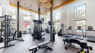 Located at 4181 Davie Rd in Davie, Florida, this fitness center offers a generously sized, single-floor exercise space designed to accommodate a wide range of workout styles and fitness goals. The facility is characterized by soaring double-height ceilings, large floor-to-ceiling windows, and an open layout that allows ample natural light to fill the space while providing outdoor views of the surrounding lush landscaping and pool area.

The main exercise floor features rubber flooring throughout and is organized into distinct training zones. The cardio area is outfitted with treadmills, stair climbers, stationary bikes, air bikes, and rowing machines, several of which are positioned beneath wall-mounted flat-screen televisions. The strength training section includes a multi-functional cable machine, a full dumbbell rack, a Smith machine, and additional cable resistance equipment.

A dedicated functional training alcove, set beneath warm pendant ring lighting and a richly toned ceiling, houses a pull-up rig, punching bag, kettlebells, medicine balls, stability balls, and a tire for functional and high-intensity workouts. Large-format athletic photography panels serve as striking visual accents throughout. Structural columns and a neutral color palette of white and dark gray create a modern, professional aesthetic. The facility is well-ventilated and thoughtfully arranged to support both individual training and group exercise sessions.