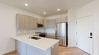 This thoughtfully designed single-floor residence showcases a clean, modern aesthetic carried consistently throughout its interior spaces. The open-concept kitchen serves as a central highlight, featuring flat-front cabinetry in a warm taupe finish accented with gold hardware, a light quartz peninsula with an undermount sink, and a full suite of stainless steel appliances including a bottom-freezer refrigerator, built-in microwave, and electric range. A soft gray subway tile backsplash adds texture while complementing the neutral color palette. Light hardwood flooring flows seamlessly from the kitchen into the living areas, enhancing the sense of openness and continuity. The living space benefits from large windows and a glass-panel exterior door that invite abundant natural light, creating a bright and airy atmosphere. A dedicated entry area features a smart lock for secure, keyless access. The bathroom is finished with matching taupe cabinetry, gold drawer pulls, a white quartz countertop, a modern chrome faucet, and a large vanity mirror. Recessed lighting and flush-mount fixtures provide consistent illumination throughout. The overall design emphasizes clean lines, neutral tones, and quality finishes, resulting in a cohesive and contemporary living environment.