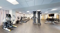 This fitness center offers a clean, functional environment designed to accommodate a wide range of exercise needs. The space features a durable speckled rubber flooring throughout, providing both safety and comfort during workouts. The drop ceiling with recessed panel lighting ensures bright, even illumination across the entire floor.

The cardio area is equipped with multiple treadmills and a stationary bike, arranged in a row along one wall accented by striped draping. A cable crossover machine takes center stage in the room, offering versatile strength training options for users of all fitness levels. Additional strength and functional training equipment, including a rowing machine, can be found along the perimeter of the space.

Large wall mirrors extend along one side of the room, enhancing the sense of openness and allowing users to monitor their form during exercise. The layout is thoughtfully organized to maximize usable floor space while maintaining clear pathways between equipment. Doorways visible at the rear of the space suggest access to additional amenity areas. Overall, the fitness center presents a well-maintained, professionally outfitted facility suitable for both casual users and dedicated fitness enthusiasts.