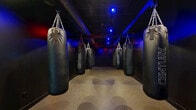 Located at 3201 First Ave in Columbus, Georgia, this purpose-built boxing bag room delivers an immersive training environment designed for serious athletic conditioning. The space is laid out in a long, narrow corridor configuration, with a row of Century heavy punching bags suspended from heavy-duty chain rigging on both sides, allowing multiple athletes to train simultaneously. The exposed ceiling infrastructure is painted black, lending an industrial aesthetic that complements the room's high-energy purpose. Dramatic colored lighting — a striking combination of blue and red spotlights — is mounted overhead, creating an atmospheric and motivating ambiance throughout the space. The walls are finished entirely in matte black, reinforcing the focused, no-distraction environment. Durable rubberized flooring runs the full length of the room, providing the traction and impact absorption necessary for boxing and combat sports training. At the far end, an open area offers additional space for warm-up, cooldown, or footwork drills. A fire extinguisher and safety equipment are properly positioned along the wall. The overall design is intentional and functional, prioritizing an intense training atmosphere while maintaining safety and accessibility for users of all skill levels.