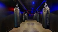 Located at 3201 First Ave in Columbus, Georgia, this purpose-built boxing bag room delivers an immersive training environment designed for serious athletic conditioning. The space is laid out in a long, narrow corridor configuration, with a row of Century heavy punching bags suspended from heavy-duty chain rigging on both sides, allowing multiple athletes to train simultaneously. The exposed ceiling infrastructure is painted black, lending an industrial aesthetic that complements the room's high-energy purpose. Dramatic colored lighting — a striking combination of blue and red spotlights — is mounted overhead, creating an atmospheric and motivating ambiance throughout the space. The walls are finished entirely in matte black, reinforcing the focused, no-distraction environment. Durable rubberized flooring runs the full length of the room, providing the traction and impact absorption necessary for boxing and combat sports training. At the far end, an open area offers additional space for warm-up, cooldown, or footwork drills. A fire extinguisher and safety equipment are properly positioned along the wall. The overall design is intentional and functional, prioritizing an intense training atmosphere while maintaining safety and accessibility for users of all skill levels.