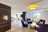 Located at 101 E Camino Real in the heart of Boca Raton, Florida, this elegantly appointed single-floor suite offers a seamlessly open layout that exudes contemporary luxury. The expansive open-plan interior is defined by light travertine flooring that flows throughout, complemented by crisp white ceilings adorned with bold, statement drum pendant lights in striking gold and yellow tones that illuminate the space with a warm, inviting glow. Distinct seating areas are thoughtfully arranged throughout, featuring deep navy blue accent chairs grouped around a polished gold spherical coffee table, alongside cream-toned lounge chairs that add contrast and sophistication. Dark, richly grained architectural columns with integrated gold accents serve as striking vertical design elements, anchoring the open floor plan. A full wet bar and kitchen area showcases warm wood cabinetry, marble-look countertops, a large mirror backsplash, and professional-grade stainless steel appliances, adjacent to a formal dining area with a sleek dark table. A media wall clad in natural wood paneling houses a large flat-screen display. Floor-to-ceiling steel-framed windows and sliding glass doors open to a balcony, offering sweeping elevated views of the Boca Raton skyline and beyond, flooding the interior with abundant natural light.
