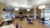 This single-floor dining space is thoughtfully designed to accommodate a variety of seating arrangements within an open, generously sized layout. The room features light wood-look flooring throughout, complemented by warm taupe walls that create a comfortable and inviting atmosphere. A drop tile ceiling hosts a series of large, cylindrical drum pendant lights that provide bright, even illumination across the dining area, while smaller black pendant fixtures accent booth and counter zones.

Seating options include freestanding tables paired with burgundy upholstered chairs with chrome frames, as well as built-in banquette booths along the perimeter walls, upholstered in a neutral woven fabric with coordinating leather backs. Framed photographic artwork and large canvas prints of natural landscapes are displayed throughout, adding visual interest and a calm, nature-inspired aesthetic.

A dedicated nook along one wall features dark cabinetry with a light countertop beneath windows overlooking greenery, decorated with fresh flowers and plants. Adjacent to this area, a classic upright piano with a padded bench serves as a charming focal point alongside additional seating, suggesting the space is used for community gatherings and entertainment. Large windows along one side bring in natural light, further enhancing the open, welcoming character of the room.