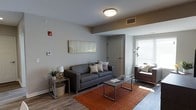 This single-floor space offers a clean, contemporary design that balances comfort and functionality. The living area serves as the centerpiece of the space, featuring warm hardwood-style flooring that grounds the room with a sense of warmth and continuity. Neutral gray walls provide a versatile backdrop, complemented by a flush-mount ceiling light that ensures even, practical illumination throughout.

The living room is thoughtfully arranged with a sleek, gray upholstered sofa accented by decorative throw pillows, paired alongside a brown accent chair positioned near a window that invites plentiful natural light. A rectangular metal-framed coffee table anchors the seating area, resting atop a rich, burnt-orange area rug that adds a bold pop of color and texture to the space.

Additional details include a side table with a table lamp and a small potted plant, adding a touch of greenery and homeliness. Wall art above the sofa contributes a subtle artistic element to the overall aesthetic. A visible entryway door and wall-mounted thermostat suggest a well-organized, functional layout. The space presents itself as a bright, move-in-ready environment with modern finishes and a welcoming ambiance suited for everyday living.