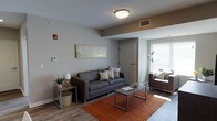 This single-floor space offers a clean, contemporary design that balances comfort and functionality. The living area serves as the centerpiece of the space, featuring warm hardwood-style flooring that grounds the room with a sense of warmth and continuity. Neutral gray walls provide a versatile backdrop, complemented by a flush-mount ceiling light that ensures even, practical illumination throughout.

The living room is thoughtfully arranged with a sleek, gray upholstered sofa accented by decorative throw pillows, paired alongside a brown accent chair positioned near a window that invites plentiful natural light. A rectangular metal-framed coffee table anchors the seating area, resting atop a rich, burnt-orange area rug that adds a bold pop of color and texture to the space.

Additional details include a side table with a table lamp and a small potted plant, adding a touch of greenery and homeliness. Wall art above the sofa contributes a subtle artistic element to the overall aesthetic. A visible entryway door and wall-mounted thermostat suggest a well-organized, functional layout. The space presents itself as a bright, move-in-ready environment with modern finishes and a welcoming ambiance suited for everyday living.