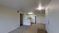 This single-floor space showcases a practical, open-concept design that seamlessly connects the living area and kitchen. The living space is finished with wall-to-wall carpeting in a neutral tone, creating a cohesive and understated aesthetic throughout. A half-wall partition defines the kitchen without fully separating it from the main living area, maintaining an open and airy feel.

The kitchen is equipped with white cabinetry arranged along the upper and lower walls, providing ample storage. A white refrigerator and a range are included among the appliances, and a fluorescent light fixture above the kitchen workspace ensures well-lit meal preparation. The kitchen floor transitions to a hard surface, distinguishing it from the carpeted living area.

Adjacent to the kitchen, a hallway leads to additional areas of the unit. A set of bifold closet doors is visible along one wall, offering convenient built-in storage. Recessed lighting and a flush ceiling fixture provide ambient illumination throughout the space. The walls are finished in a clean, light neutral shade, and HVAC vents are integrated into both the ceiling and lower wall areas, ensuring climate comfort throughout the unit.