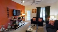 This single-floor residence offers a thoughtfully designed living space that combines bold style with everyday comfort. The living room immediately draws attention with its striking burnt-orange accent wall, which serves as a dramatic backdrop for the wall-mounted flat-screen television and decorative shelving unit. The warm tones are complemented by soft, neutral carpeting that runs throughout, creating a cozy and inviting atmosphere. A ceiling fan with integrated lighting provides both airflow and ambient illumination, enhancing the room's comfort year-round. Natural light flows generously into the space through a large window dressed with dark curtains, as well as through a glass-paneled exterior door that opens to the outdoors, offering a seamless connection to the outside. The living area is furnished with a dark sofa accented by patterned throw pillows in warm hues, and a glass-topped coffee table that adds a modern touch to the eclectic décor. A console-style media stand with open shelving provides practical storage while contributing to the room's layered aesthetic. The overall ambiance is warm, lively, and well-appointed, making it a welcoming and functional living environment.