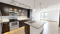 This thoughtfully designed one-bedroom, one-bathroom unit offers a cohesive modern aesthetic throughout its single-floor layout. The open-concept kitchen and living area serves as the heart of the space, anchored by a granite-topped center island with a stainless steel sink and pendant lighting above. Dark espresso cabinetry with brushed hardware lines the kitchen walls, complemented by a white subway tile backsplash, granite countertops, and a full suite of stainless steel appliances including a refrigerator, range, and built-in microwave. Track lighting illuminates the kitchen and living area, which flows seamlessly toward a large floor-to-ceiling window that frames sweeping city and river views. Light hardwood flooring runs throughout the entire unit, lending warmth and continuity. The bedroom is bright and airy, featuring a glass balcony door and an additional window with panoramic views of the surrounding cityscape and a notable suspension bridge. The bathroom is well-appointed with dark cabinetry, a granite vanity, a framed mirror, a soaking tub with tiled surround, and continued hardwood flooring. A closet and hallway round out the layout, completing this efficiently planned and stylishly finished space.