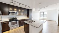 This thoughtfully designed one-bedroom, one-bathroom unit offers a cohesive modern aesthetic throughout its single-floor layout. The open-concept kitchen and living area serves as the heart of the space, anchored by a granite-topped center island with a stainless steel sink and pendant lighting above. Dark espresso cabinetry with brushed hardware lines the kitchen walls, complemented by a white subway tile backsplash, granite countertops, and a full suite of stainless steel appliances including a refrigerator, range, and built-in microwave. Track lighting illuminates the kitchen and living area, which flows seamlessly toward a large floor-to-ceiling window that frames sweeping city and river views. Light hardwood flooring runs throughout the entire unit, lending warmth and continuity. The bedroom is bright and airy, featuring a glass balcony door and an additional window with panoramic views of the surrounding cityscape and a notable suspension bridge. The bathroom is well-appointed with dark cabinetry, a granite vanity, a framed mirror, a soaking tub with tiled surround, and continued hardwood flooring. A closet and hallway round out the layout, completing this efficiently planned and stylishly finished space.