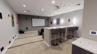 This private movie theater is designed to deliver an immersive cinematic experience within a well-appointed, single-level space. The viewing area is anchored by a large projection screen mounted on a neutral gray accent wall, served by a ceiling-mounted projector and a surround sound speaker system integrated throughout the room. Seating is arranged in two tiered rows of black leather reclining chairs, each equipped with cup holders, ensuring clear sightlines from every seat. The raised rear platform provides an elevated second row for unobstructed viewing.

The décor takes on a distinct theme, with bold Art Deco-style Marvel superhero posters—featuring Spider-Man, Thor, Iron Man, Captain America, and others—displayed prominently along the walls. Wall sconces provide ambient accent lighting, complementing the recessed ceiling lights that illuminate the space. Neutral carpet tiles and light-toned baseboards lend a clean, polished aesthetic throughout.

A built-in snack bar adds significant functionality to the space, featuring a granite countertop, dark cabinetry, a stainless steel microwave, and a classic popcorn machine. Adjacent bar-height seating along a counter offers an alternative viewing area. Potted plants add touches of greenery, completing this well-equipped, themed entertainment venue.