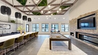 This expansive single-floor community lounge offers a sophisticated blend of modern design and inviting comfort. The space is immediately distinguished by its soaring ceilings, adorned with a striking geometric wood beam structure interwoven with lush living greenery and suspended Edison-style pendant lights, creating a dramatic yet organic overhead focal point.

The living area is anchored by a full-height exposed brick accent wall featuring a wide linear fireplace and a large wall-mounted television, providing a warm and stylish centerpiece. Generous sectional seating arranged on a patterned area rug creates an inviting lounge zone, while a full-size billiard table positioned at the heart of the room adds a recreational dimension. Large black-framed windows line the perimeter, flooding the interior with abundant natural light and offering views to the surrounding exterior.

The kitchen area showcases a lengthy waterfall-edge marble-look countertop island with seating for multiple guests, complemented by sculptural pendant fixtures and open shelving with a patterned tile backsplash. A cozy nook near the entrance provides semi-private booth-style seating beneath pendant lights. Wide-format tile flooring flows throughout, unifying the various zones of this well-appointed, multifunctional gathering space.