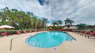 Located at 1900 Bella Vista Way in Port St. Lucie, Florida, this community pool area offers a resort-style outdoor experience designed for relaxation and recreation. The centerpiece is a generously sized, freeform swimming pool with crystal-clear turquoise water, featuring a gradual entry point marked at 3 feet, making it accessible for a range of swimmers. The pool is surrounded by a broad, light-colored concrete sun deck outfitted with rows of vibrant red lounge chairs, providing ample space for sunbathing and leisure. A shaded umbrella station is also available poolside for those seeking relief from the Florida sun. The atmosphere is elevated by the lush tropical landscaping that frames the entire area, including tall, swaying palm trees and mature green foliage that create a sense of privacy and seclusion. The natural backdrop of dense trees and neighboring residential structures can be seen beyond the perimeter, reinforcing the community setting. Open to the sky above, the pool deck benefits from abundant natural light, with the surrounding greenery lending a serene, tropical ambiance characteristic of South Florida living. This outdoor amenity space serves as an inviting communal retreat within the Bella Vista Way community.