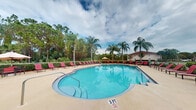 Located at 1900 Bella Vista Way in Port St. Lucie, Florida, this community pool area offers a resort-style outdoor experience designed for relaxation and recreation. The centerpiece is a generously sized, freeform swimming pool with crystal-clear turquoise water, featuring a gradual entry point marked at 3 feet, making it accessible for a range of swimmers. The pool is surrounded by a broad, light-colored concrete sun deck outfitted with rows of vibrant red lounge chairs, providing ample space for sunbathing and leisure. A shaded umbrella station is also available poolside for those seeking relief from the Florida sun. The atmosphere is elevated by the lush tropical landscaping that frames the entire area, including tall, swaying palm trees and mature green foliage that create a sense of privacy and seclusion. The natural backdrop of dense trees and neighboring residential structures can be seen beyond the perimeter, reinforcing the community setting. Open to the sky above, the pool deck benefits from abundant natural light, with the surrounding greenery lending a serene, tropical ambiance characteristic of South Florida living. This outdoor amenity space serves as an inviting communal retreat within the Bella Vista Way community.