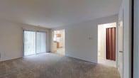 This single-floor residential unit offers a clean, functional layout designed for comfortable everyday living. The main living area is finished with neutral wall-to-wall carpeting that adds warmth underfoot, and the bright white walls create a light, open atmosphere throughout. A large sliding glass door fitted with vertical blinds lines one wall of the living space, allowing for generous natural light and providing direct access to an outdoor area.

The living area connects seamlessly to the kitchen through an open doorway, offering a convenient and accessible flow between spaces. The kitchen features warm wooden cabinetry paired with light countertops and tile flooring, complemented by a white refrigerator and appliances that give the room a clean, practical feel. The tile flooring in the kitchen provides a durable contrast to the carpeted living area.

A separate doorway off the main living space leads to additional rooms, including what appears to be a hallway or secondary area, suggesting a thoughtful floor plan that balances open common spaces with more private zones. The overall ambiance of the unit is bright, neutral, and move-in ready, with a simple, understated aesthetic that provides a versatile backdrop for a variety of interior styles.