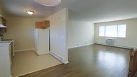 This single-floor unit features an open-concept layout that seamlessly connects the kitchen and living areas. The kitchen is partially defined by a dividing wall and includes warm-toned wood cabinetry and a full-size white refrigerator, with carpeted flooring in that zone transitioning into rich hardwood floors that extend throughout the living area. The living space is generously sized, bathed in natural light from a window along the far wall. Baseboard heating units run along the walls in both areas, providing efficient climate control throughout. The neutral, light beige wall tones create a clean and versatile backdrop. A ceiling fan is mounted overhead near the kitchen, adding to the unit's functionality. The overall ambiance is straightforward and practical, offering an open, airy feel well-suited for everyday living.
