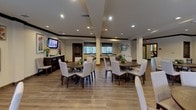 This single-floor communal dining space is designed to accommodate a large number of guests across a generously sized, open floor plan. The room features warm wood-look flooring throughout, lending a welcoming and cohesive aesthetic to the entire space. Multiple round dining tables, each surrounded by upholstered cream-toned chairs, are arranged throughout the main area, encouraging social interaction and comfortable group dining. A long communal-style table with bench seating offers an additional dining option along one side of the room.

Recessed lighting is installed throughout the ceiling, providing consistent, well-distributed illumination across the space. Exposed beam detailing along the ceiling adds architectural character and visual depth. Warm yellow-toned walls are accented with decorative artwork, sunburst wall mirrors, and decorative shelving, contributing to the inviting ambiance.

Natural light filters in through multiple windows dressed with blinds and patterned curtains, offering views to the exterior. A dedicated alcove area contains a smaller, more intimate round dining table surrounded by upholstered chairs, ideal for private gatherings. A dark wood sideboard with decorative accessories serves as a functional and stylish accent piece near the entrance area. A wall-mounted television provides entertainment within the dining environment.