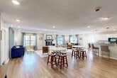 Located at 1011 Gadd Rd in Chattanooga, TN, this single-floor space offers an open-concept layout that seamlessly integrates the living room, dining area, and kitchen into one cohesive, functional environment. Warm hardwood-style flooring runs throughout, tying the spaces together and contributing to a welcoming ambiance.

The living room is a standout feature, anchored by a whitewashed brick fireplace that serves as an eye-catching focal point. Multiple large windows dressed with light gray curtains flood the area with natural light and provide views of the surrounding greenery. Recessed lighting adds to the bright, airy atmosphere.

The dining area comfortably accommodates multiple seating arrangements with round high-top tables and bar-height stools, making it ideal for both casual gatherings and everyday use. A wall-mounted television and additional console table enhance the room's functionality.

The kitchen is well-equipped with white cabinetry, granite countertops, a breakfast bar with bar seating, and stainless steel appliances. A pendant light fixture above the bar adds a charming finishing touch, while a window overlooking the outdoors brings in additional natural light. The overall design is clean, modern, and highly practical.