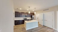 This single-floor unit offers a thoughtfully designed layout with a neutral color palette and contemporary finishes throughout. The kitchen serves as a central highlight, featuring rich dark-stained shaker-style cabinetry extending to the ceiling for ample storage, light quartz countertops, and a full suite of stainless steel appliances including an electric range, built-in microwave, and a French door refrigerator. A spacious center island with an undermount sink and pendant lighting above provides both additional workspace and a natural gathering point. Wood-style luxury vinyl plank flooring runs through the kitchen and main living areas, lending warmth and durability. The living space is open and accessible, connecting directly to the bedroom areas. Bedrooms feature plush carpeting and large windows that allow natural light while offering views of the surrounding building exterior. Ceiling fans are present for added comfort. Both bathrooms are finished with dark vanity cabinetry, light countertops, wall-mounted storage cabinets, and wood-style flooring consistent with the rest of the unit. A dedicated laundry area is also present within the unit, adding practical convenience to the overall floor plan.