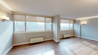 This single-floor residence offers a well-designed layout that balances open living spaces with functional supporting rooms. The expansive main living area is defined by wide-plank hardwood flooring that runs throughout, neutral gray walls, and large windows fitted with roller shades that frame elevated city skyline views. Baseboard heating units line the perimeter beneath the windows, and wall-mounted sconces provide warm accent lighting. Crown molding trims the ceiling in the living and dining areas, adding a refined architectural detail. A chandelier anchors the dining space, which connects seamlessly to the kitchen through a pass-through window opening.

The galley-style kitchen is efficiently organized with dark espresso cabinetry on both sides, light granite countertops, a subway tile backsplash, and stainless steel appliances including a range, microwave, and dishwasher. Beadboard wainscoting on the lower walls adds a classic touch.

The bathroom is finished with white subway tile walls accented by a decorative mosaic tile border, a granite-topped vanity with an undermount sink, dark wood cabinetry, and a wall-mounted sconce beside a framed mirror. Chrome fixtures and grab bars complete the clean, polished look throughout.