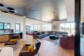 Located at 1401 Third Ave S in Birmingham, Alabama, this single-floor communal living space offers a generously sized, open-plan lounge designed with a bold, contemporary aesthetic. The room's most striking architectural feature is its dramatic deep-blue painted tray ceiling, accented with sweeping gold-toned linear details and anchored by a sculptural mid-century-inspired chandelier, creating an eye-catching focal point throughout the space.

Large floor-to-ceiling windows line the perimeter, flooding the interior with abundant natural light and offering expansive views of the Birmingham cityscape and nearby landmarks. Light-toned wood-look flooring runs throughout, providing a warm contrast to the richly colored furnishings.

The space is thoughtfully organized into multiple distinct seating zones. One area features a large sectional sofa in slate gray, layered with teal and earth-tone accent pillows, arranged around a pair of oval coffee tables. A second lounge area showcases vibrant teal velvet chairs atop a deep crimson area rug, centered around tiered glass coffee tables beneath a vivid floral art piece. Additional leather chairs with brass frames add a sophisticated, eclectic character.

A built-in wet bar with wood cabinetry and open shelving serves as a functional amenity, complemented by a wall-mounted television framed by textured teal accent paneling. A glass-paned entry door provides a sleek, modern transition into the space.