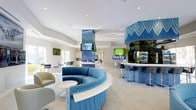 This single-floor space showcases a contemporary open-concept design with a bright, airy atmosphere enhanced by white walls, high ceilings, and recessed lighting throughout. The layout is thoughtfully arranged to encourage both socializing and relaxation.

A standout feature is the curved, circular seating arrangement upholstered in rich blue velvet, centered around a sleek round marble-top table — creating an inviting focal point within the lounge area. Adjacent to it, a curved bar station wrapped in bold blue patterned fabric with coordinating bar stools serves as a stylish social hub, complemented by mounted screens for entertainment.

A striking central column clad in a vivid blue geometric-patterned fabric rises toward the ceiling, anchoring the space visually while hosting additional display screens. The flooring consists of large-format light-toned tiles that reflect the abundant natural light streaming in through generous windows.

Further into the space, a more casual dining or activity area is visible, alongside what appears to be a kitchenette or service area in the background. Accent wall art in green and blue tones adds visual interest. The overall ambiance is energetic yet comfortable, with a cohesive color palette of blue, white, and green.