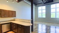 This single-floor space showcases a compelling blend of industrial and modern design. The open layout is anchored by exposed dark ceiling beams and structural steel columns, lending an authentic loft character to the interior. Polished hardwood floors run throughout, reflecting natural light and adding warmth to the space.

A built-in granite countertop with dark-stained cabinetry serves as a natural divider between the kitchen and living areas. The cabinetry features clean-lined hardware and ample storage, complementing the granite's speckled finish. Under-cabinet lighting adds a functional and aesthetic touch.

The living area benefits from a striking arched window that draws in generous amounts of natural light, offering views of the surrounding greenery. The ceiling fan above contributes to the industrial aesthetic while providing practical air circulation. The exposed ceiling structure, combining dark wood and metal elements, adds visual depth and height to the space.

Overall, the unit presents a cohesive industrial-chic aesthetic, balancing raw architectural details with refined finishes to create a functional and visually dynamic environment.