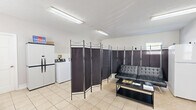 Located at 3761 S State Ave in Indianapolis, Indiana, this single-floor community facility offers a practical and welcoming environment designed to serve residents with essential shared amenities. The space is organized into distinct functional zones, separated by a decorative folding room divider that provides a degree of privacy between areas.

The laundry area is equipped with multiple commercial-grade washers and dryers arranged along the walls, all set atop light-colored ceramic tile flooring that extends throughout the entire space. A fire extinguisher and posted safety notices reflect the facility's attention to safety standards. Fluorescent overhead lighting ensures the area is well-lit throughout.

A separate resource and lounge area features computer workstations with office chairs, a printer, and a shared full-size refrigerator and microwave, making it a functional hub for residents' everyday needs. A welcoming multilingual banner decorates the wall, reinforcing the inclusive character of the facility. Comfortable seating, including a futon sofa and a coffee table, provides a space for residents to wait or relax. Natural light enters through windows fitted with blinds, adding brightness to the interior. The overall atmosphere is clean, utilitarian, and community-oriented.