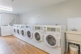 Located at 400 Parkside Dr in Waterloo, Ontario, this shared laundry facility is a well-organized, single-room space designed to serve building residents efficiently. The room features warm wood-look flooring throughout, which contrasts neatly with the crisp white walls and ceiling, creating a clean and functional environment.

The facility is equipped with a generous number of commercial-grade front-loading washing machines and top-loading dryers, all finished in white and arranged along the walls to maximize usable floor space. A dedicated row of dryers lines one wall, complete with visible ventilation ductwork routed along the ceiling and walls, ensuring proper airflow and safe operation. Additional top-load washers with utility sinks are positioned in a separate alcove, offering versatility for various laundry needs.

A built-in folding counter runs along one wall, providing a convenient surface for sorting and folding laundry. Bright fluorescent ceiling fixtures illuminate the space evenly, while a window on the far wall allows for natural light and ventilation. Safety features include a clearly marked exit door, a fire extinguisher, and a security camera. The room is accessed through a paneled white door and maintains a tidy, well-maintained appearance throughout.