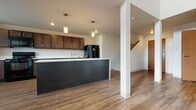 This two-story residence offers a thoughtfully designed layout that blends modern finishes with functional living spaces. The main level centers around an open-concept kitchen and living area, where rich hardwood-style flooring flows throughout. The kitchen is well-equipped with dark espresso cabinetry, white countertops, a full suite of black appliances including a range, microwave, dishwasher, and refrigerator, and a large island with a contrasting dark face panel. Pendant lighting above the island adds a contemporary touch. A half bathroom on the main level features matching dark cabinetry, a white countertop, and a large mirror. The living area benefits from soaring ceilings and a sliding glass door that opens to an outdoor space, allowing generous natural light to pour in through both the door and a high transom window above.

A carpeted staircase with a warm wood handrail leads to the upper level, where bedrooms are finished with plush carpet and windows offering exterior views. The upper-level full bathroom includes a sliding glass-enclosed shower, dark vanity cabinetry, and wood-look flooring consistent with the home's overall aesthetic. A ceiling fan in one bedroom adds comfort and functionality to the upper floor.